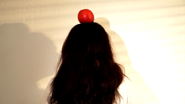 Rückansicht einer Person mit langen, dunklen Haaren vor einer hellen Wand; ein roter Apfel liegt zentral auf dem Kopf. Weiches Licht und Schatten erzeugen eine grafische Wirkung.